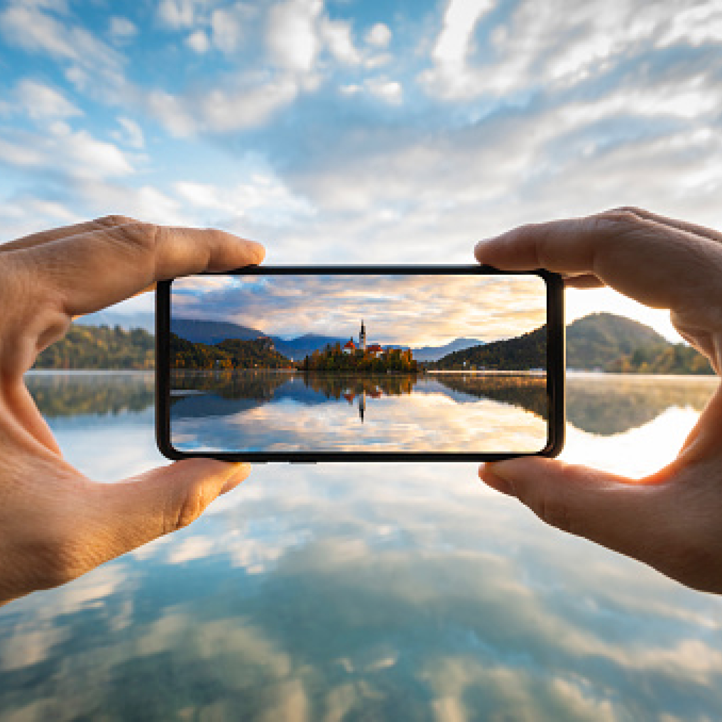 Person macht ein Landschaftsfoto an einem See mit einer kleinen Insel und Bergpanorama im Hintergrund.